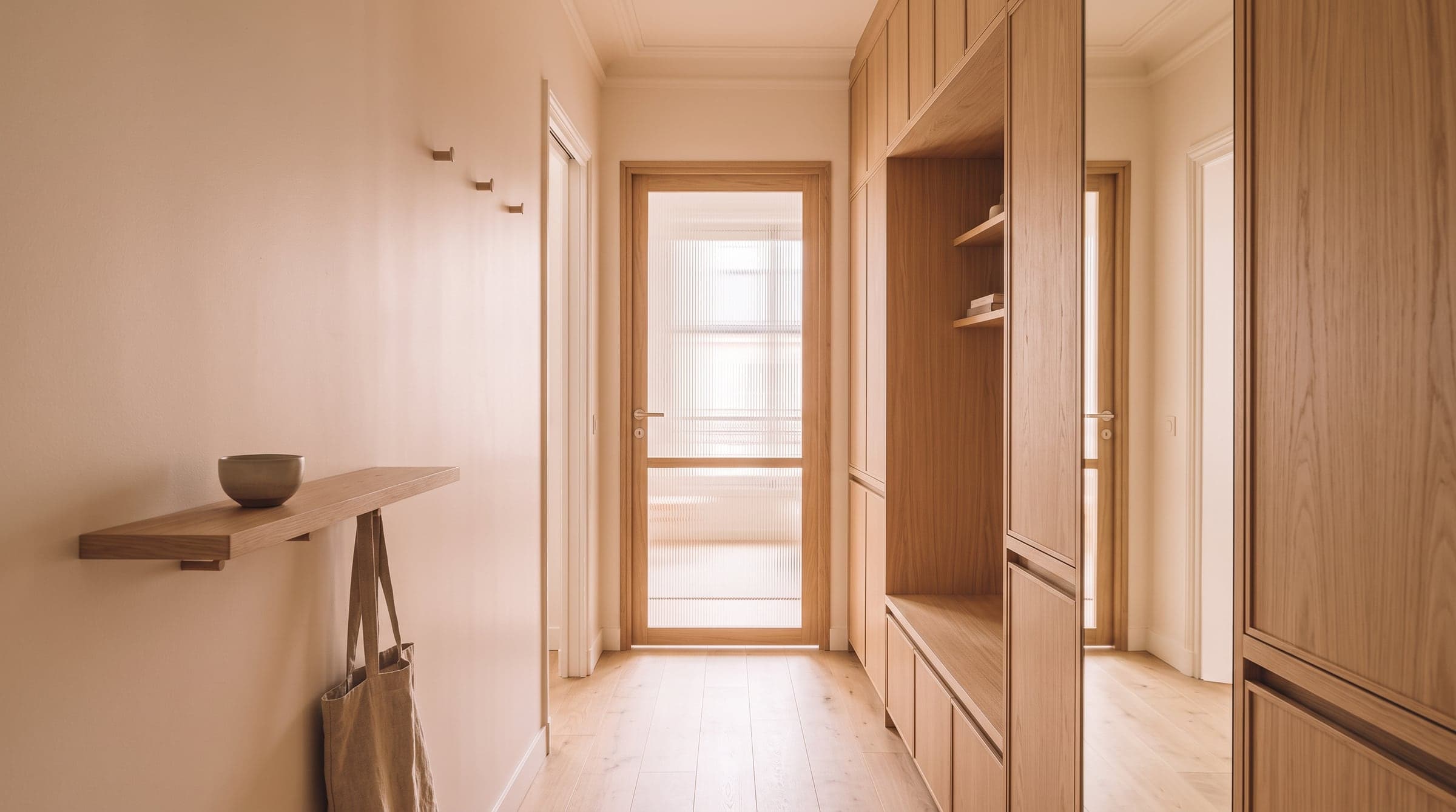 View down a hallway toward a glazed door, light reflected by a tall mirror set into the wall joinery