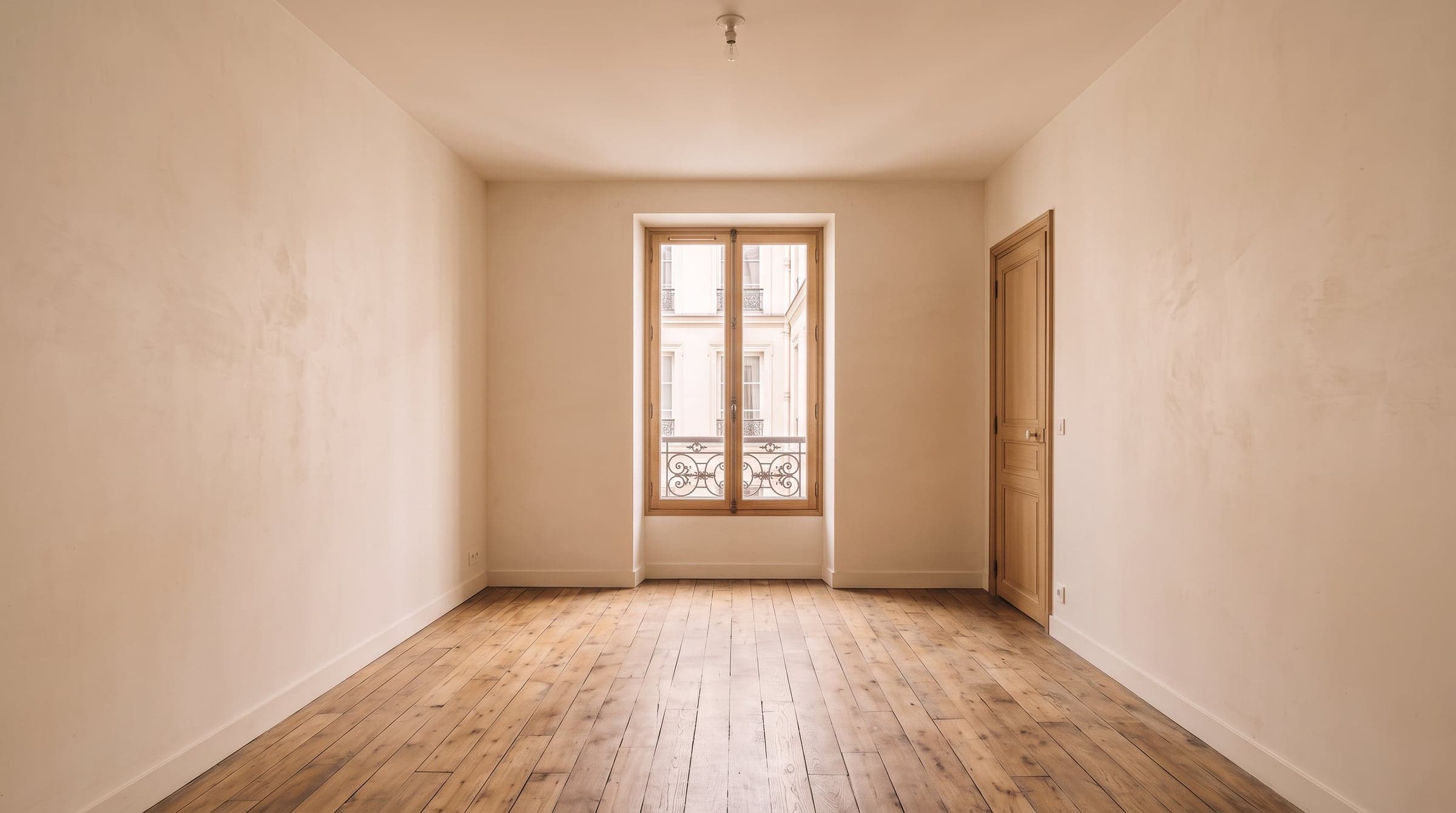 A dim, courtyard-facing Paris room with a narrow corridor door, plain plaster walls and a single small window