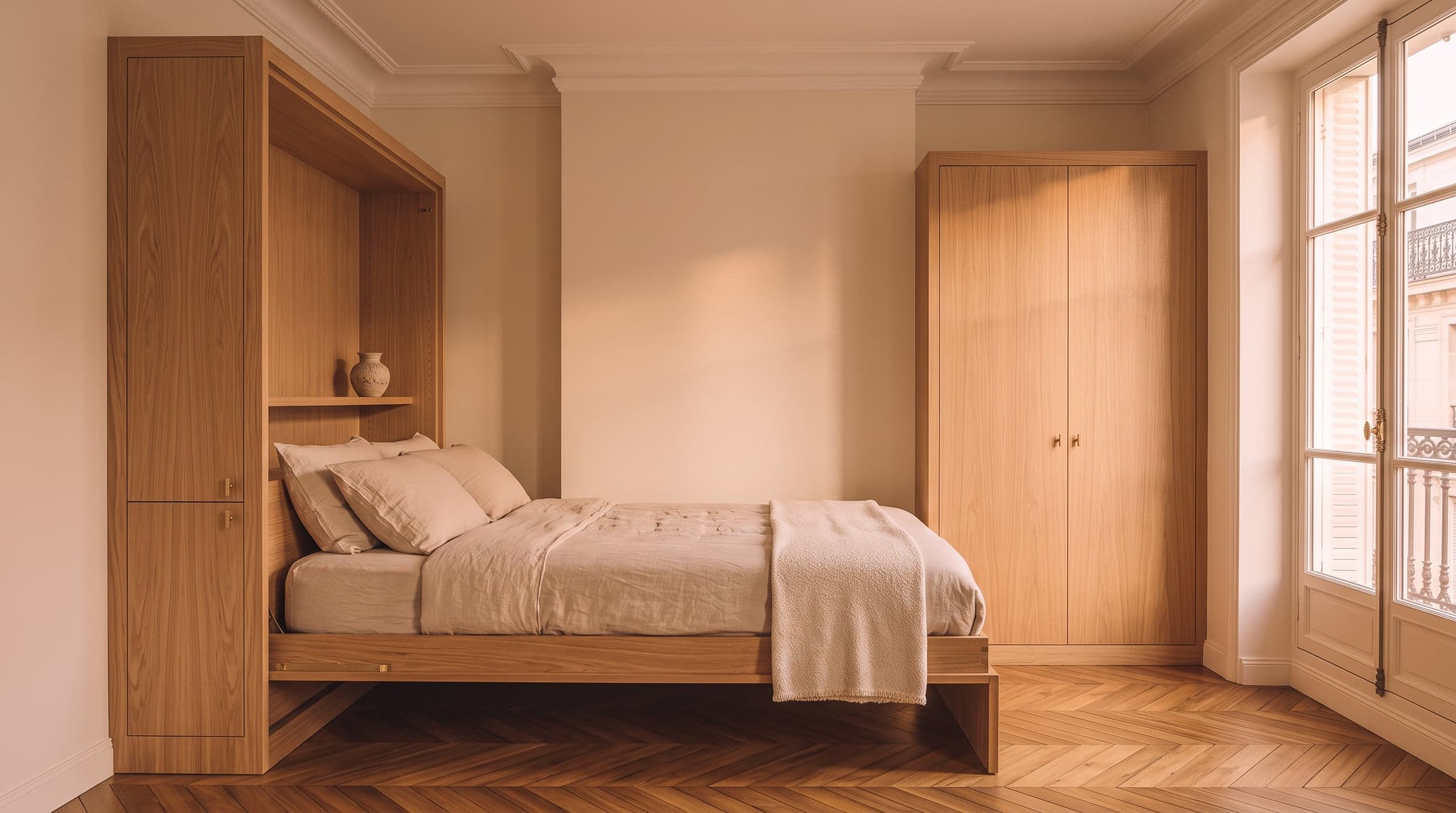 Fold-away bed lowered from a wall of joinery in a small Paris apartment, made up with linen sheets, evening light through a tall window